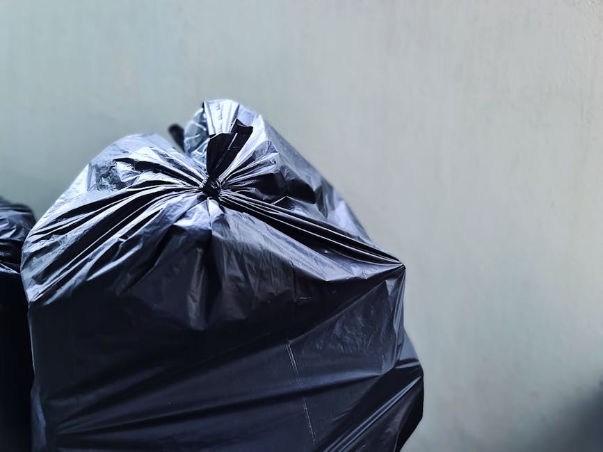 A large pile of mixed waste and trash bags accumulated around a central metal recycling container on a paved urban street. The waste consists of various cardboard boxes, paper bags, plastic packaging,
