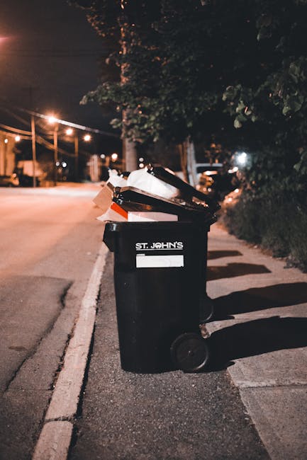 A black plastic wheelie bin labeled 'ST. JOHN'S' positioned on a residential sidewalk at night, filled with various rubbish including flattened cardboard boxes, paper, and plastic waste, some of which